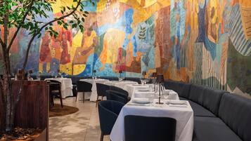 A restaurant with tables and chairs, a mural on the wall, and a tree in the corner.