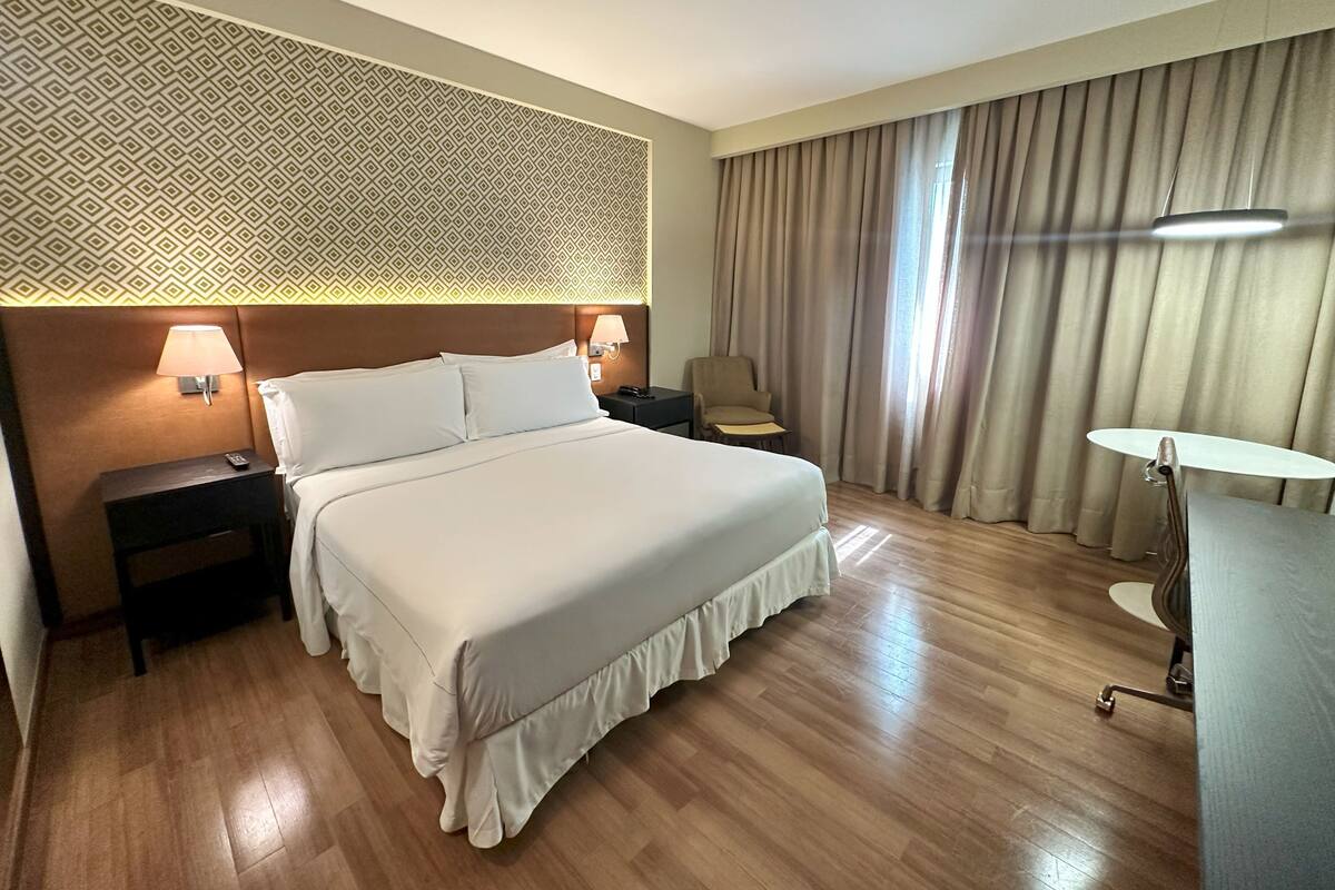 Une chambre d’hôtel avec un grand lit, deux tables de chevet avec des lampes, une chaise, un bureau avec une chaise et un mur décoré d’un motif.