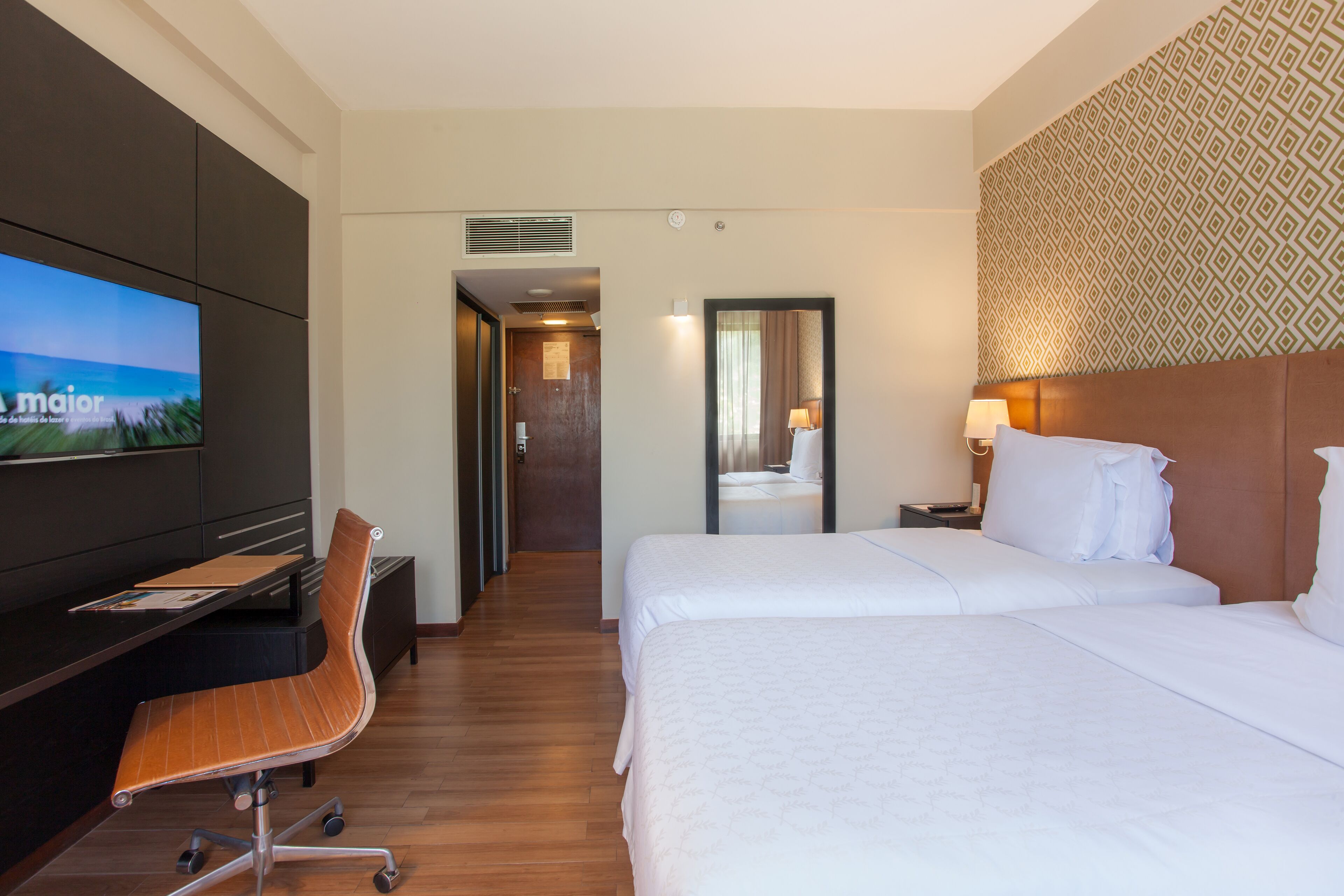 Quarto de hotel com cama, escrivaninha, cadeira, televisão e banheiro visível por uma porta aberta.