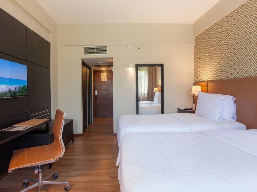 Quarto de hotel com cama, escrivaninha, cadeira, televisão e banheiro visível por uma porta aberta.