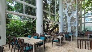 Een eethoek binnen met tafels en stoelen, grote ramen en uitzicht op groen buiten.