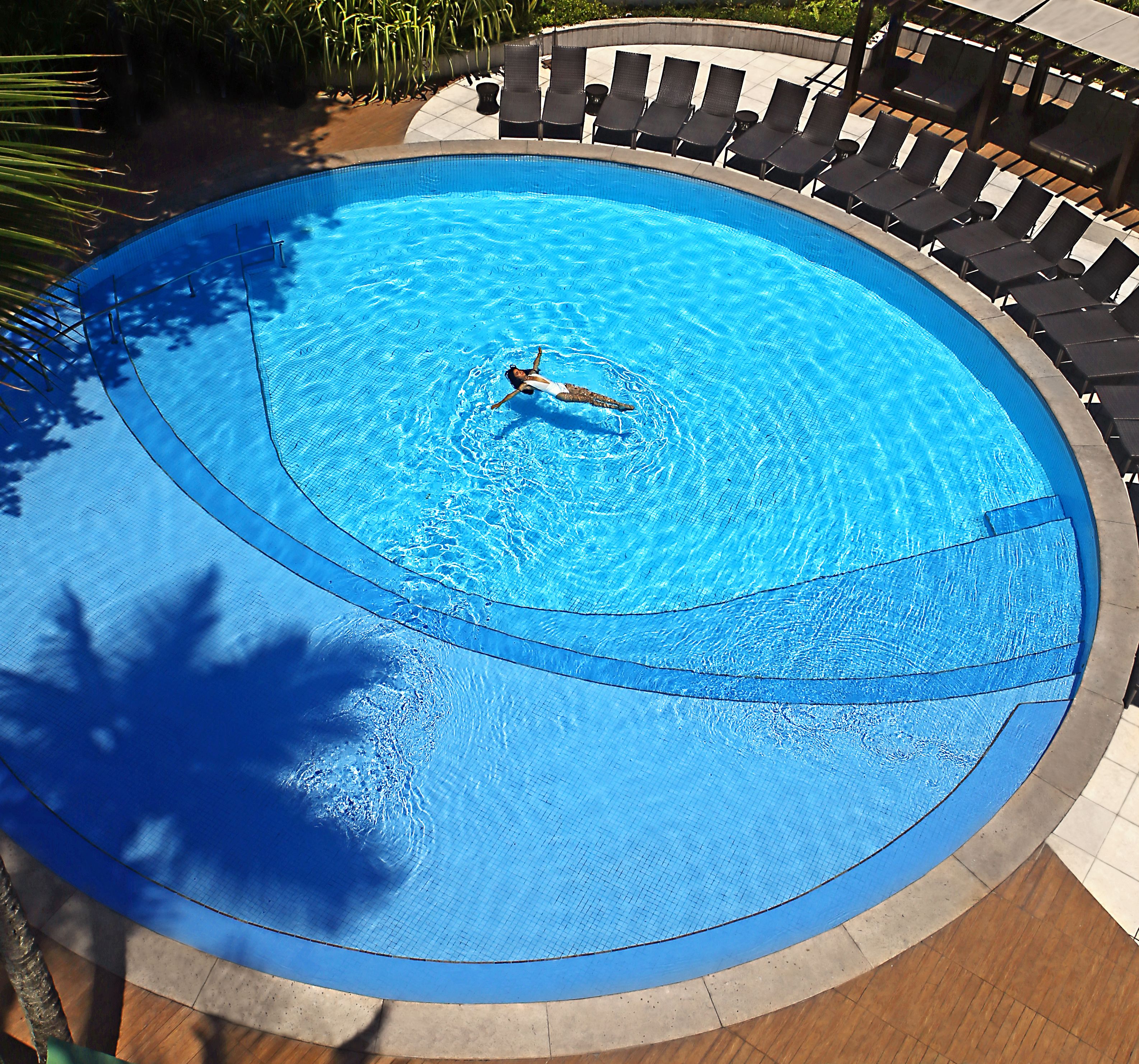A circular pool with a person swimming, surrounded by lounge chairs and a tiled deck.