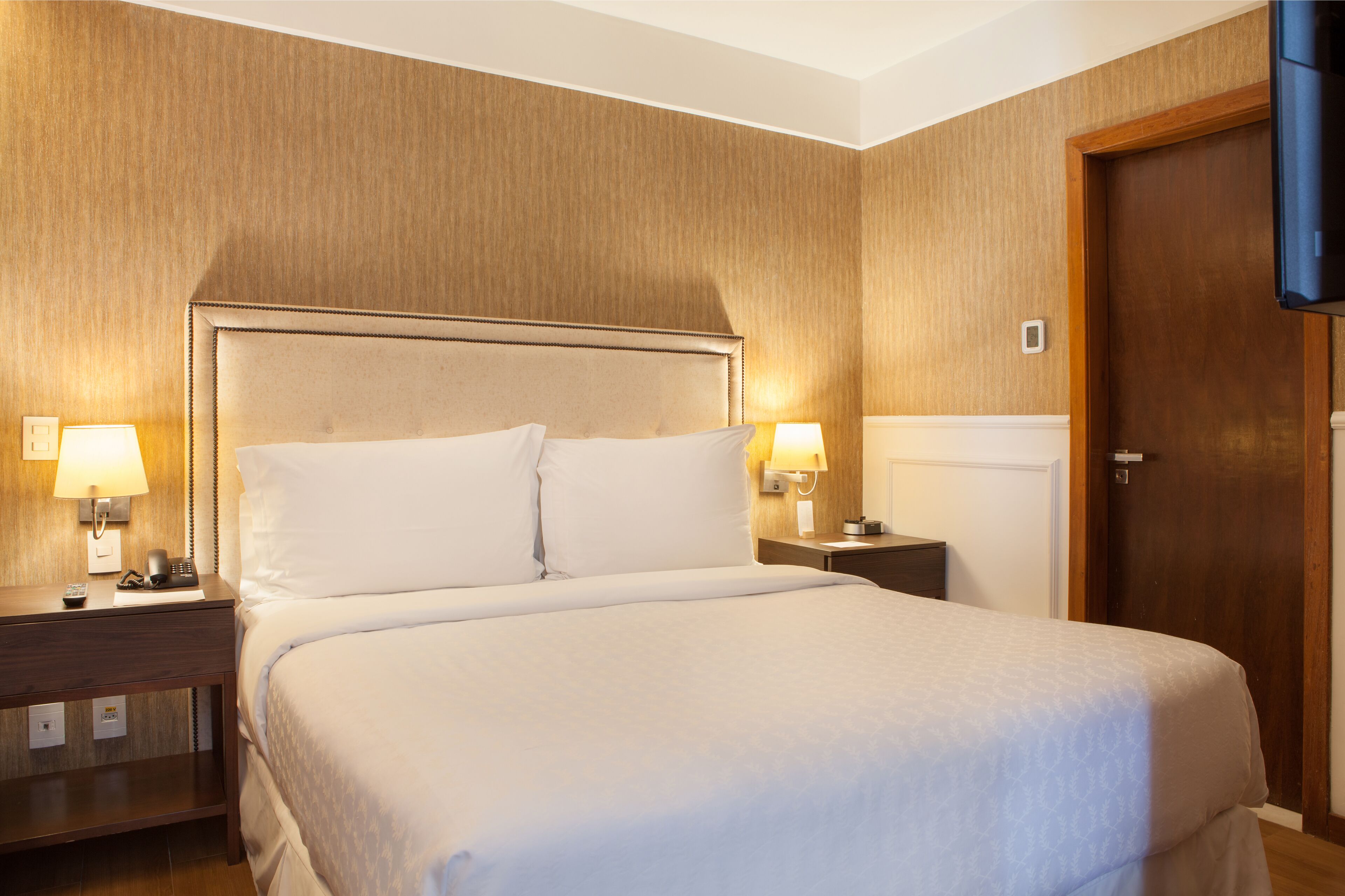 Una habitación de hotel con una cama grande, dos mesitas de noche con lámparas, una puerta de madera y un televisor de pantalla plana montado en la pared.