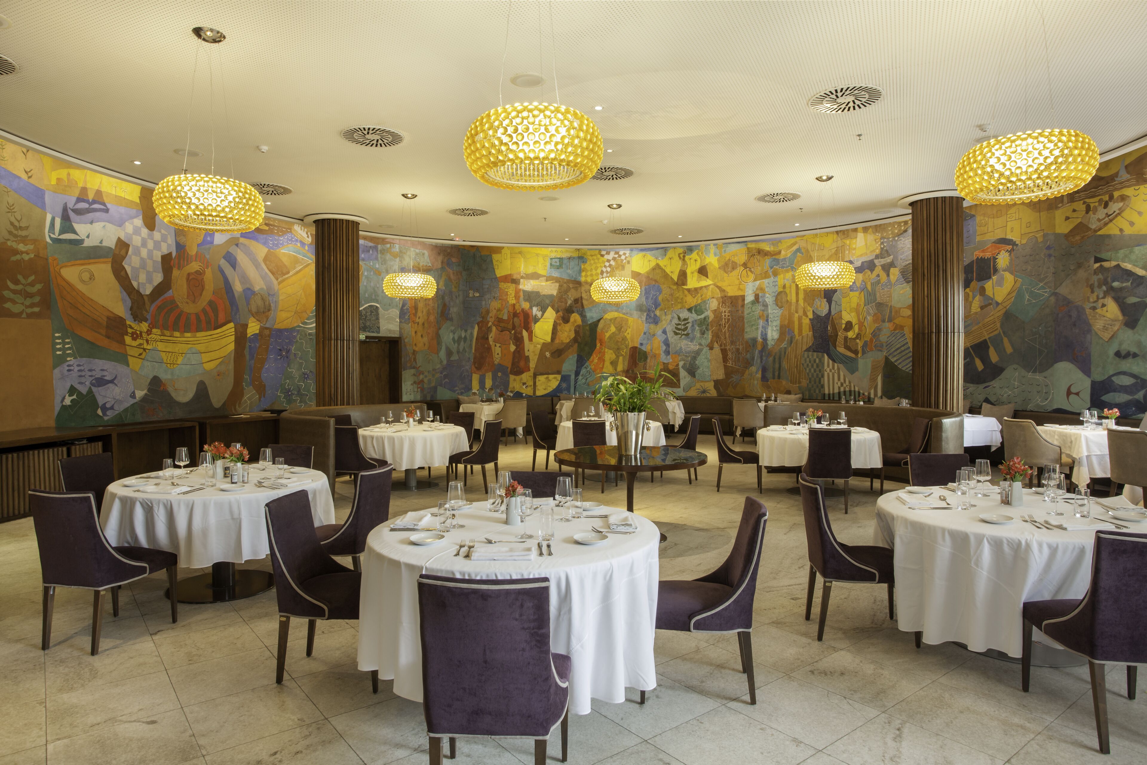 Uma área de refeições com mesas redondas, cadeiras roxas e iluminação decorativa.