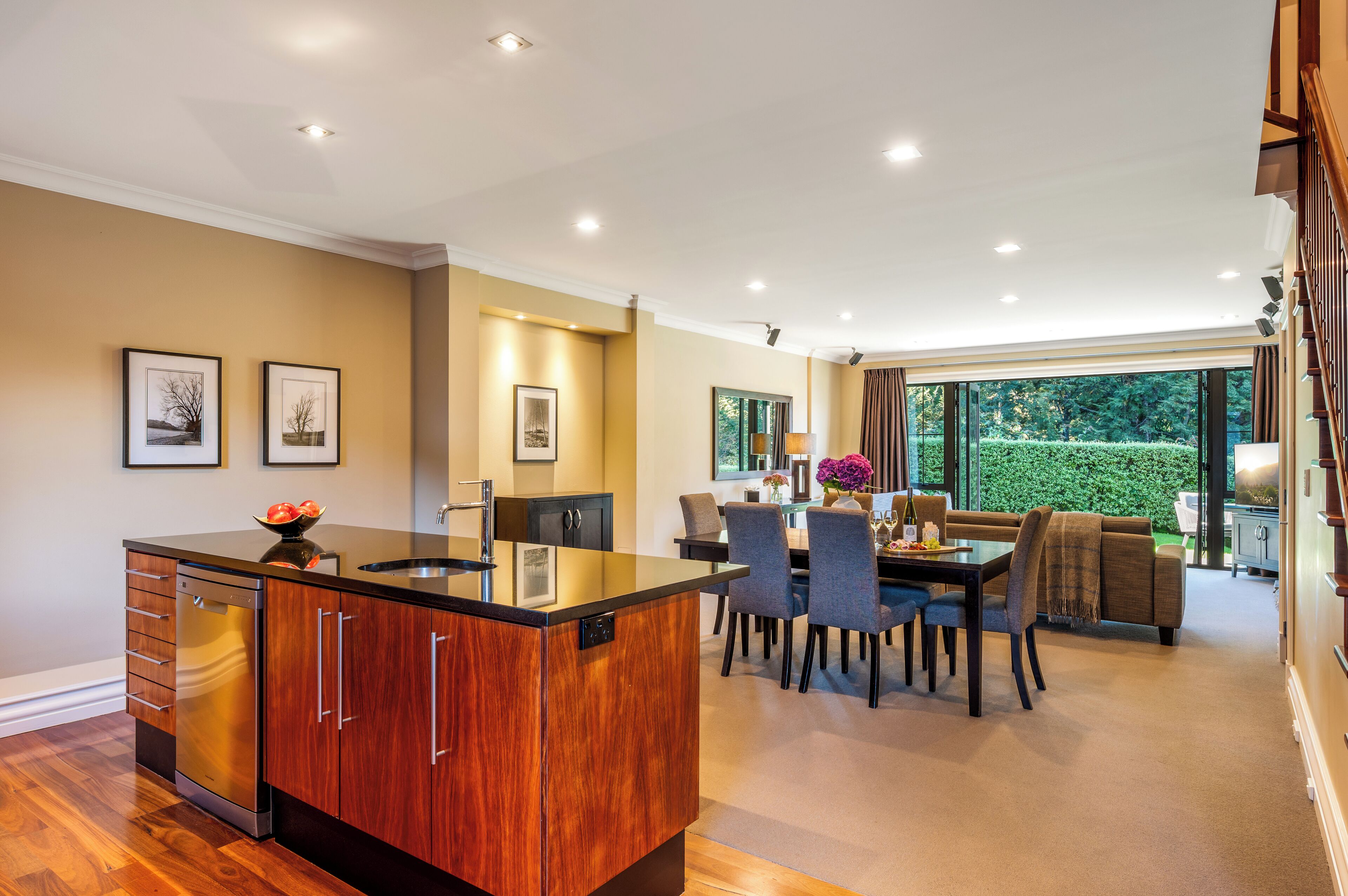 A modern kitchen with a central island, dining area with chairs and a table, and a view of a garden through large windows.