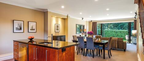 A modern kitchen with a central island, dining area with chairs and a table, and a view of a garden through large windows.