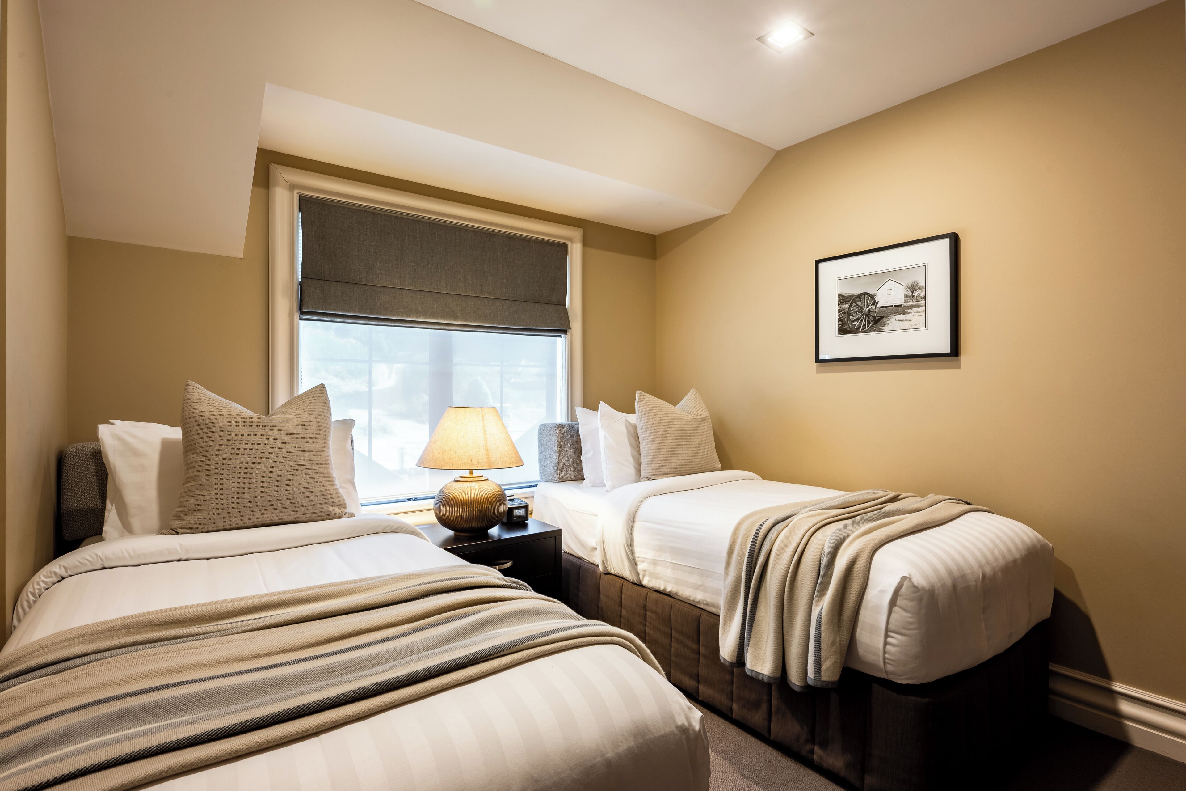 Quarto de hotel com duas camas, uma mesinha de cabeceira com abajur, uma janela com cortina e um quadro na parede.