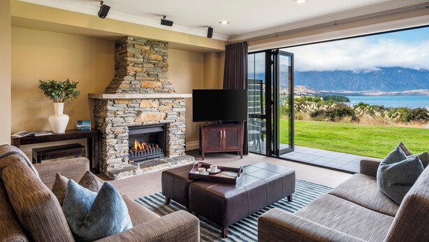 A living room with a stone fireplace, a flat-screen TV, and a large window offering a scenic view of mountains and a lake. - Platinum Queenstown Villas (Queenstown)