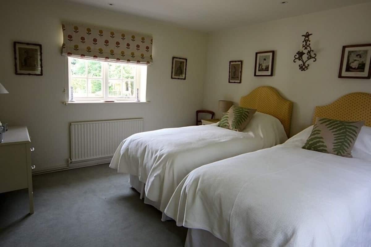 A bedroom with two beds, a window with a patterned curtain, a radiator, and several framed pictures on the walls.