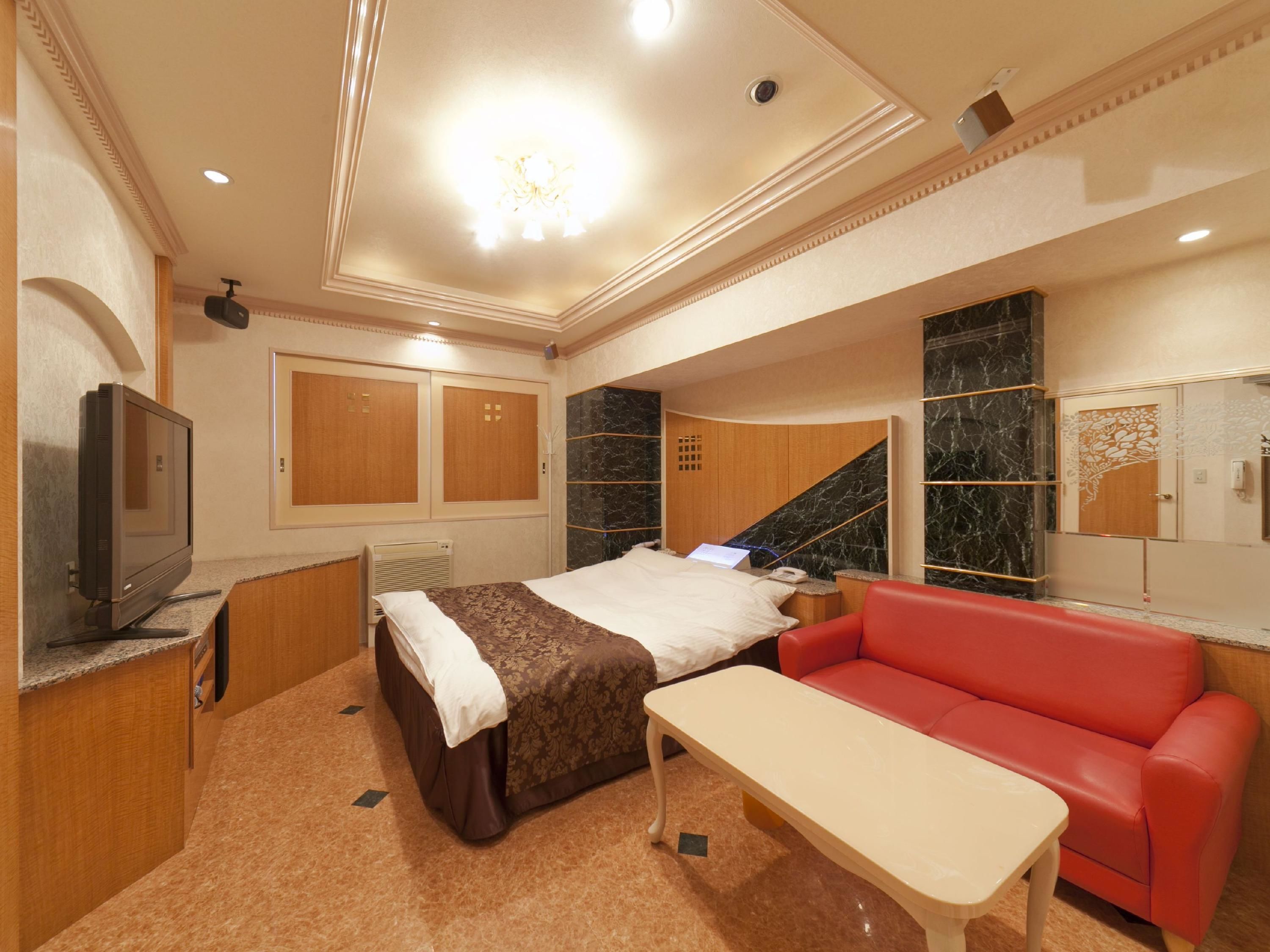 Habitación de hotel con una cama, un sofá rojo, una mesita, un televisor y un baño con pared de mármol.