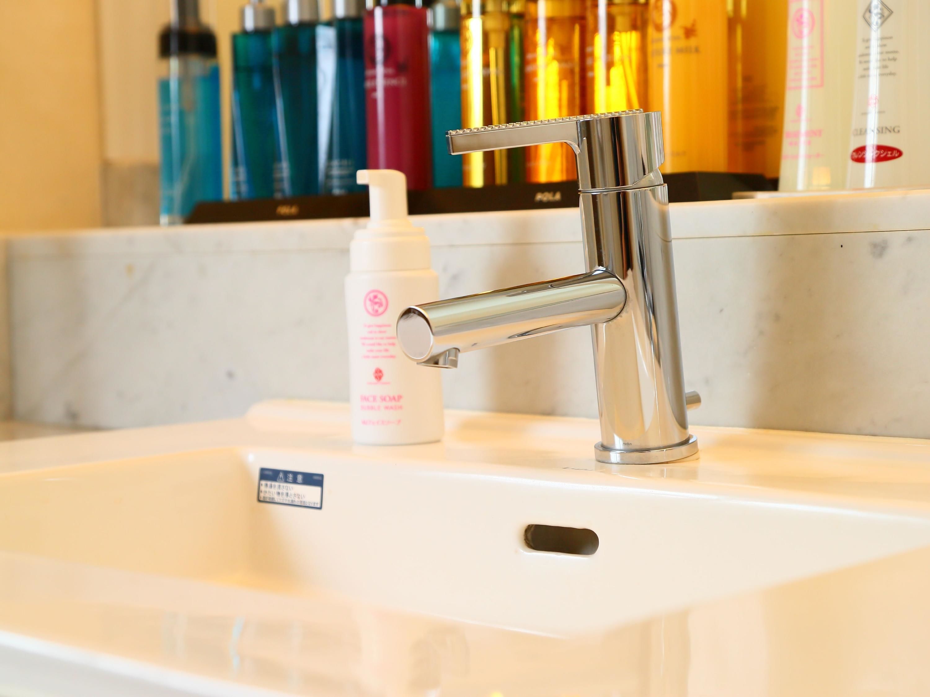 A bathroom sink with a modern faucet, a bottle of facial soap, and various toiletries in the background.