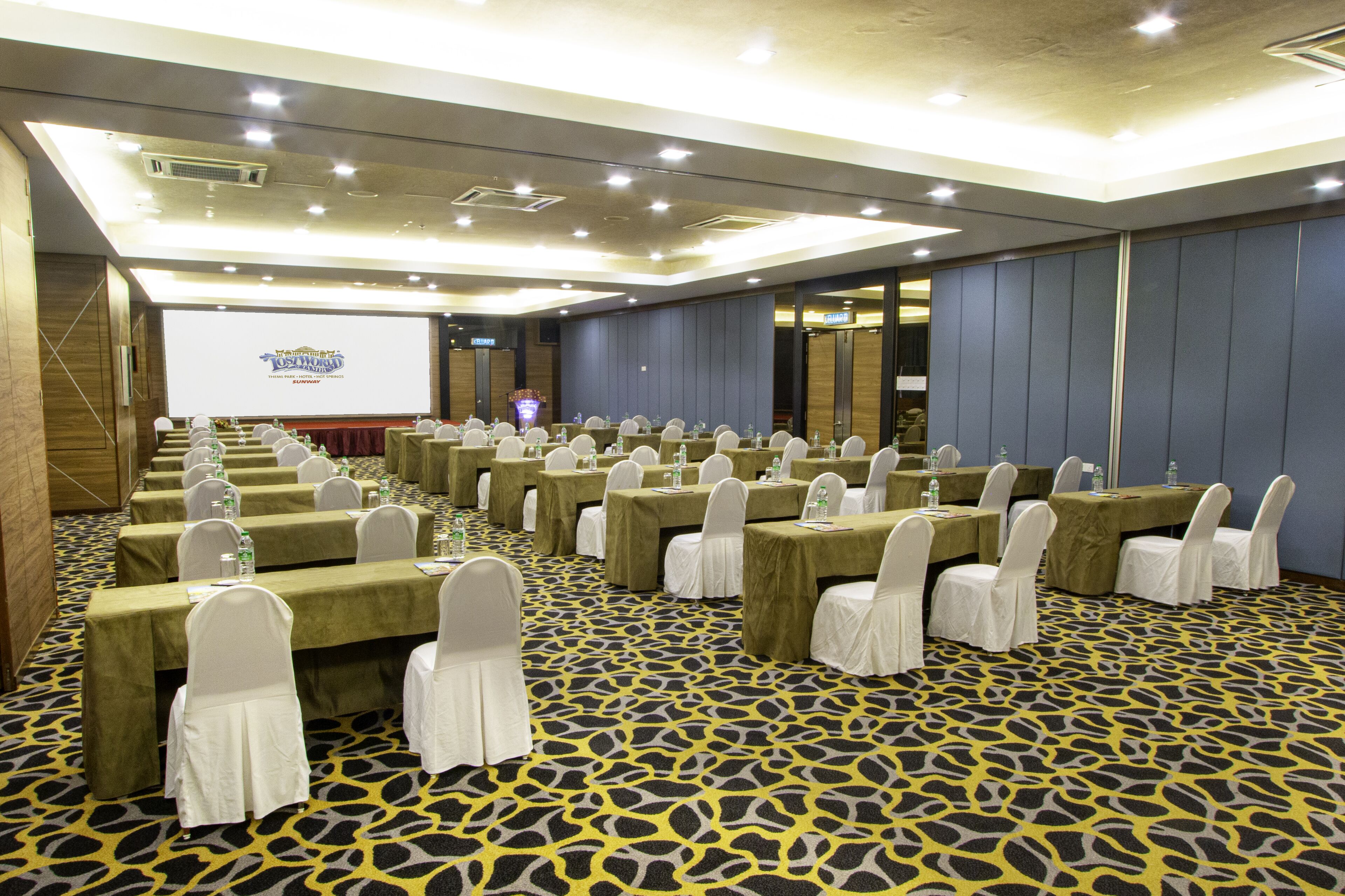 A conference room with rows of tables and chairs, a projection screen, and a patterned carpet.