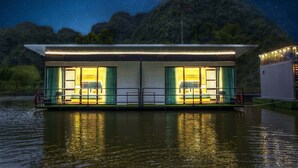 Un hotel flotante con dos habitaciones iluminadas, un balcón y vistas a un cielo estrellado y montañas.