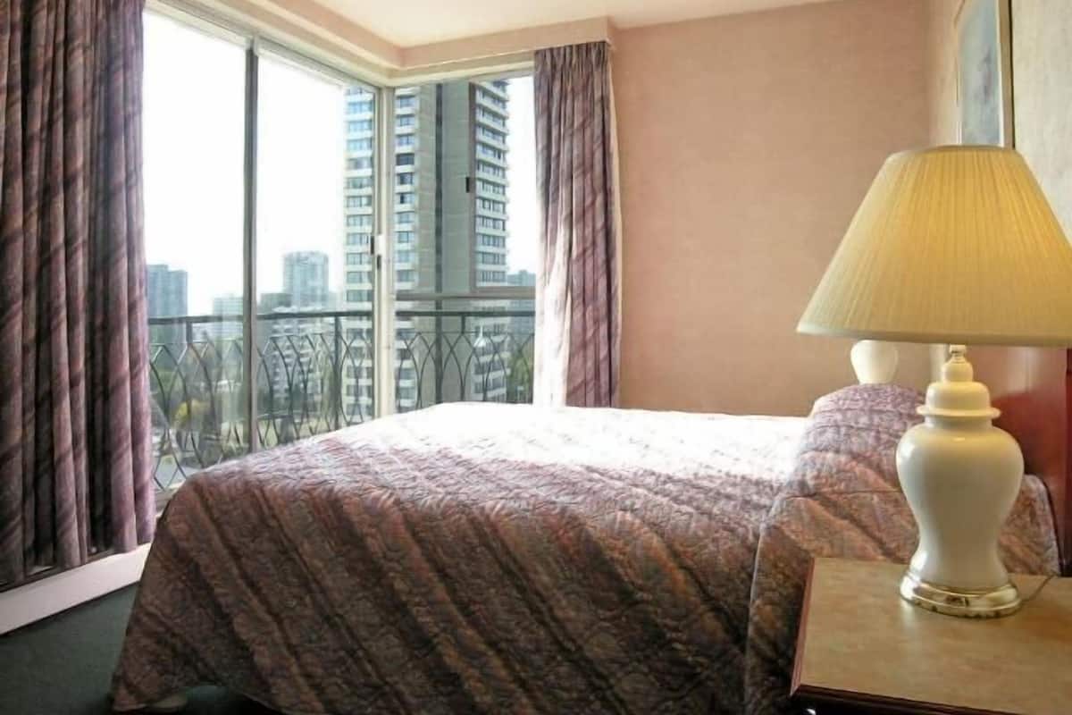 A hotel room with a large bed, a nightstand, a lamp, and a view of tall buildings through the window.