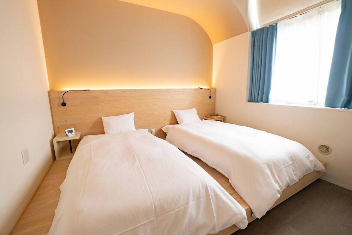 Habitación de hotel con dos camas, una mesita de noche con lámpara y una ventana con cortinas.