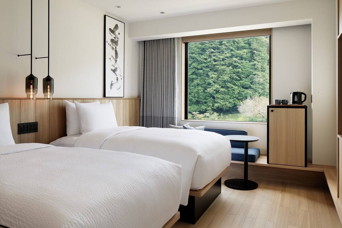 Habitación de hotel con dos camas, un ventanal grande con vista al bosque, una mesita y un televisor.