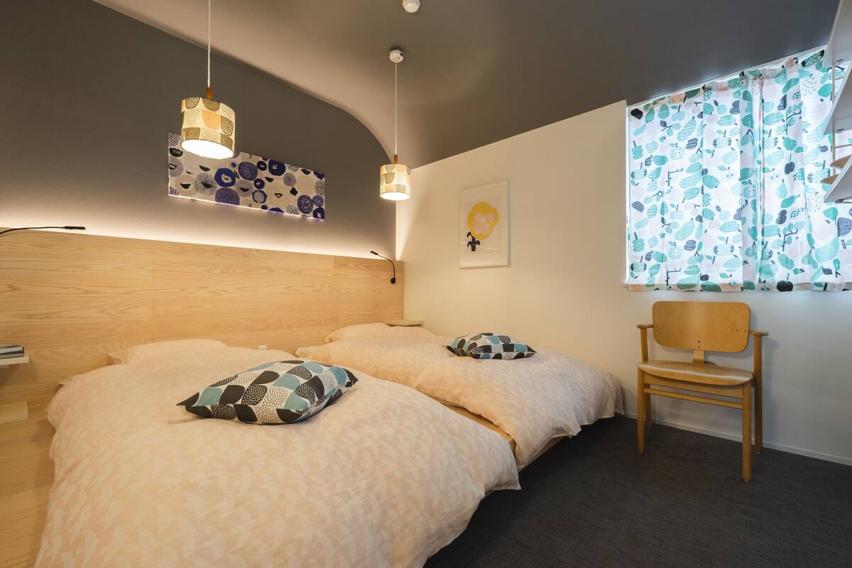 Un dormitorio con dos camas, cabecero de madera, una silla, una ventana con cortinas estampadas y una lámpara fijada a la pared.