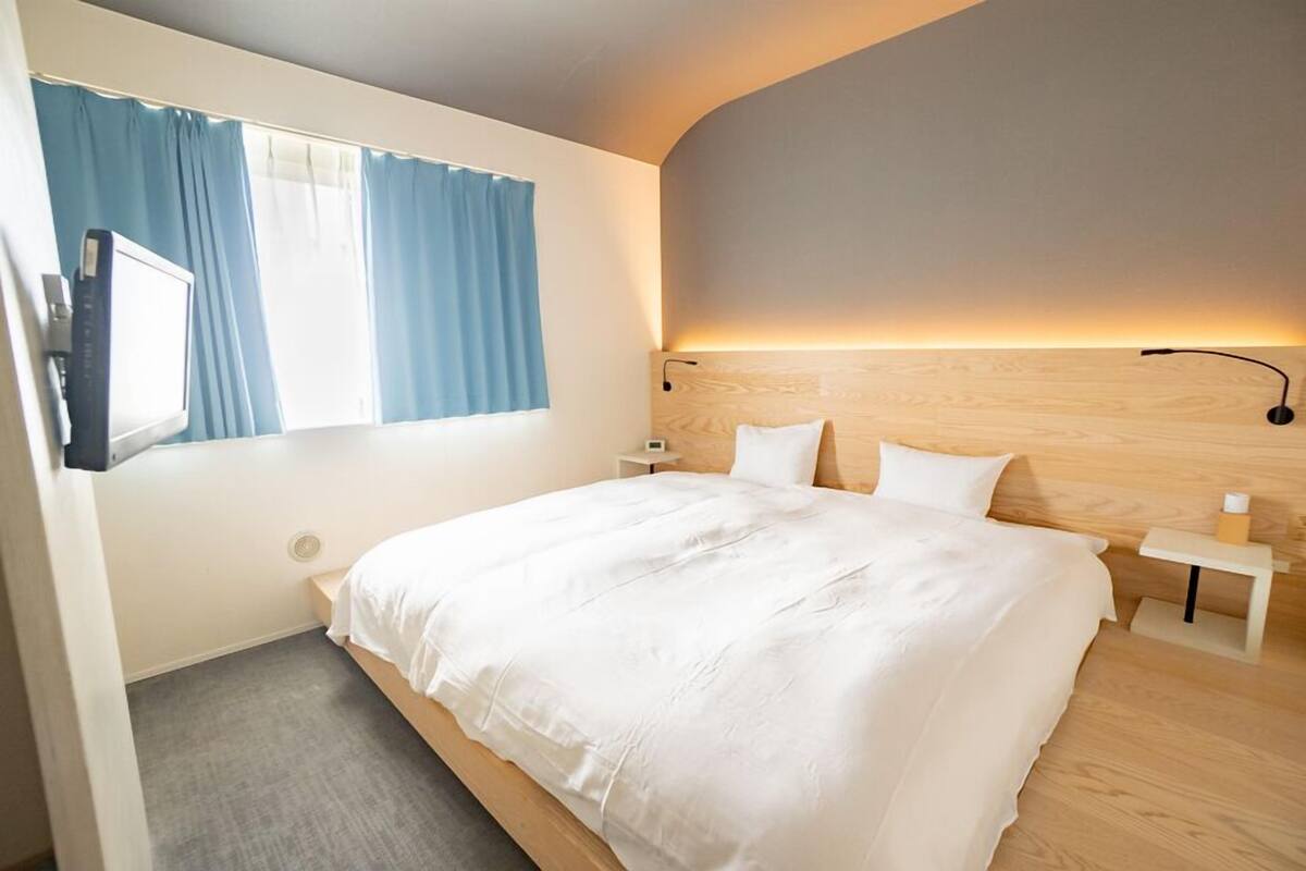Un dormitorio con cama, mesita de noche, lámpara, televisor y ventana con cortinas.
