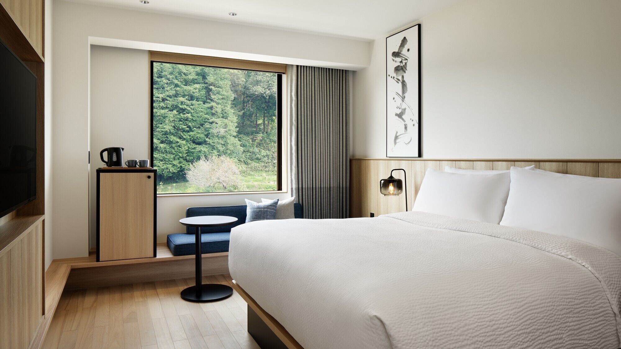 A hotel room with a large bed, a small table, a TV, a window with a view of trees, and a painting on the wall.