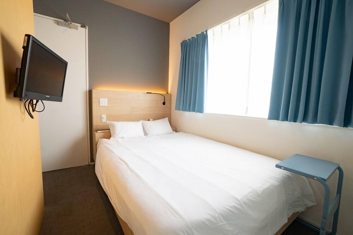 Habitación de hotel con cama, televisor montado en la pared, mesita de noche y ventana con cortinas.