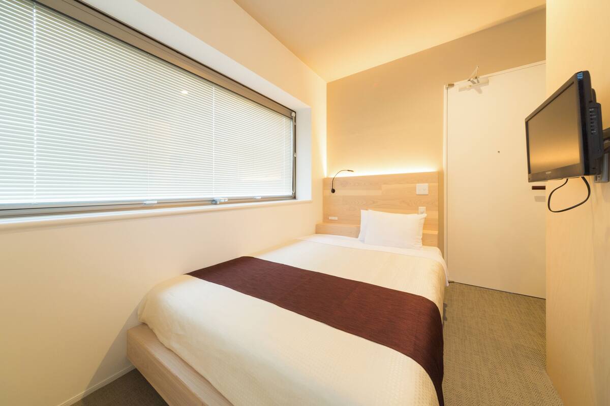 Una habitación de hotel con cama, televisión, ventana con persianas y puerta.
