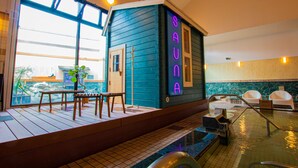 Zona de spa con una cabaña de madera que dice "SAUNA", un banco, una mesita y vistas al exterior a través de amplios ventanales.