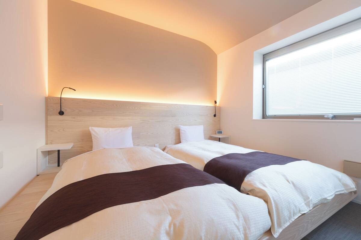 Una habitación de hotel moderna con dos camas, un cabecero de madera, mesitas de noche y una ventana con persianas.