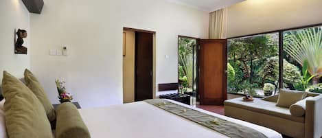 A hotel room with a large bed, a sofa, a view of greenery through a window, and a door leading to another room.