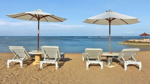 Una playa con sillas de descanso blancas y sombrillas, una mesa pequeña, y una estructura roja a lo lejos.