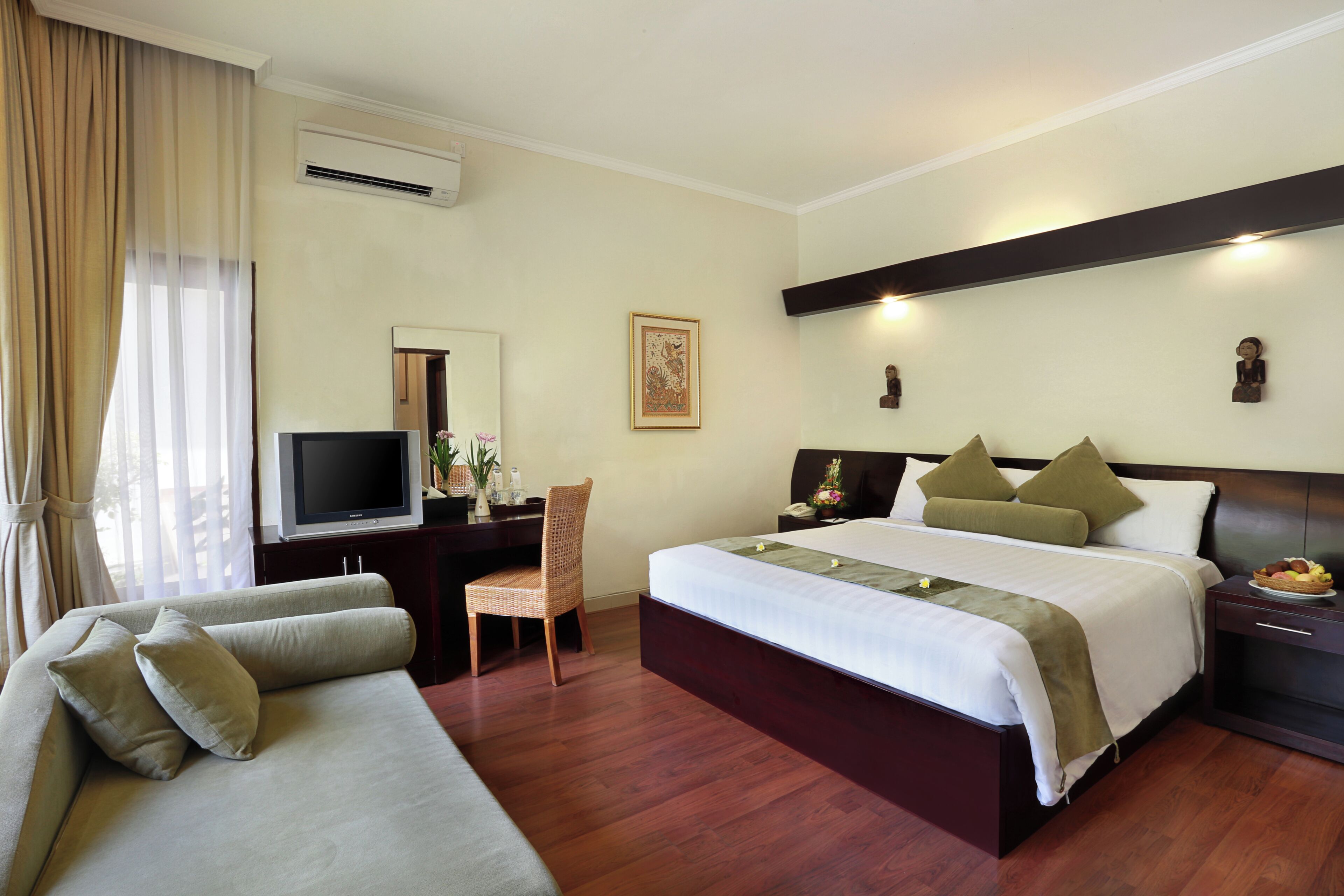 A hotel room with a bed, a sofa, a desk with a television, and a wall-mounted air conditioner.