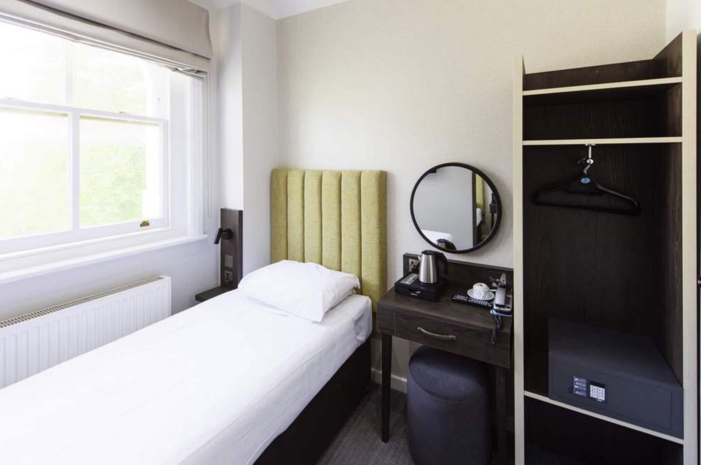 Ein Hotelzimmer mit Bett, Schreibtisch mit Wasserkocher, Spiegel, Kleiderhaken und Kleiderschrank.