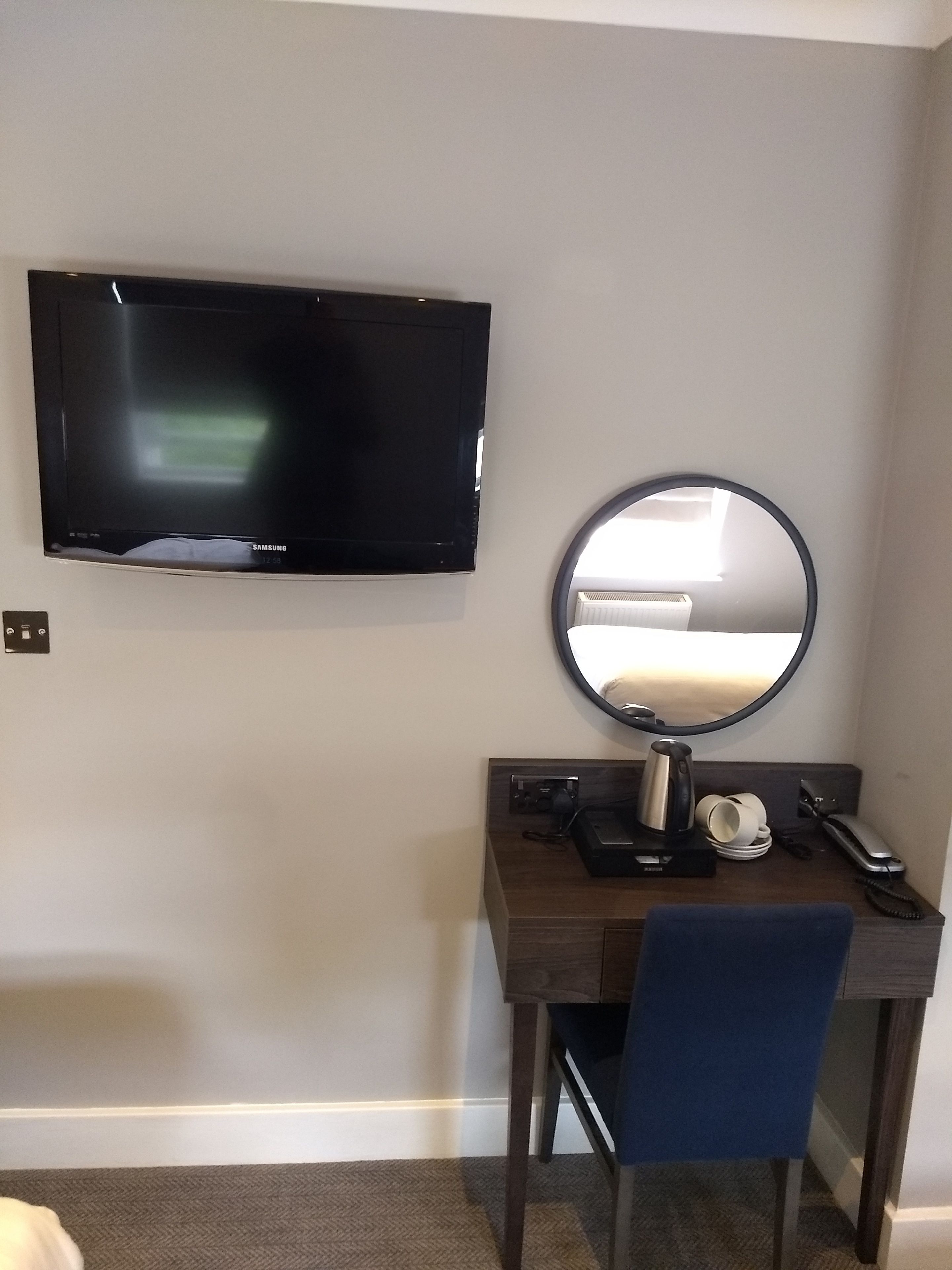 Een hotelkamer met een flatscreen televisie aan de muur, een ronde spiegel, een bureau met een stoel, en een klein tafeltje met een waterkoker en kopjes.