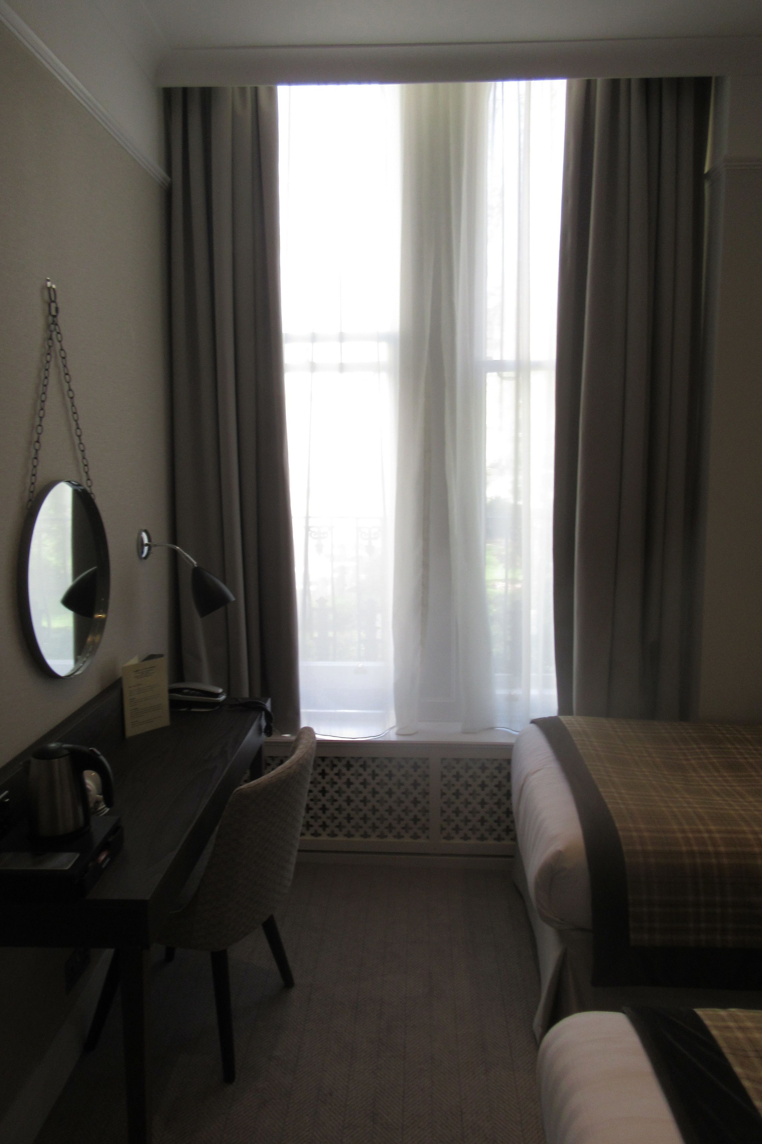 Habitación de hotel con cama, escritorio, silla, espejo y ventana con cortinas.