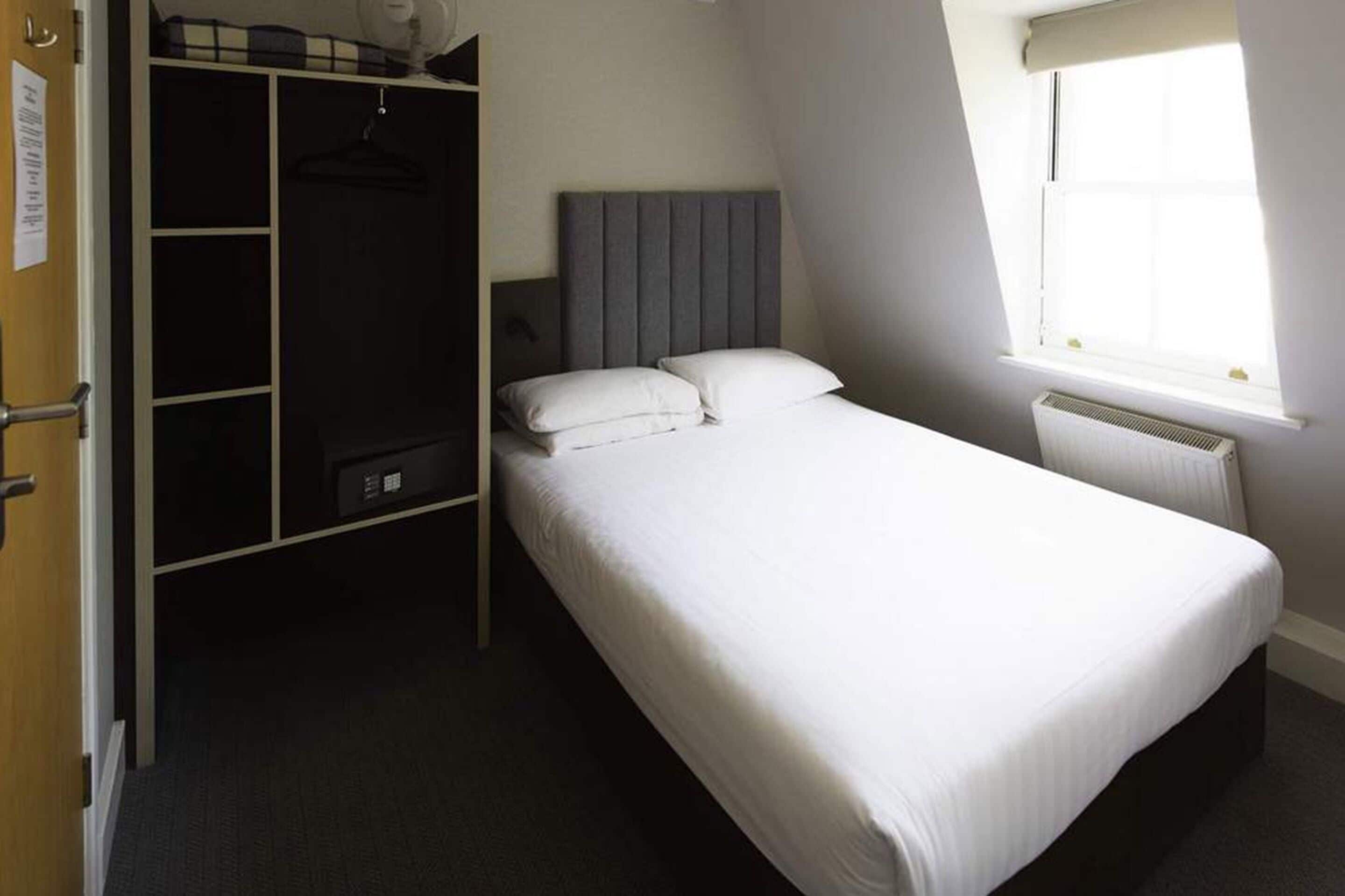 A single bed with a grey headboard, a wardrobe with a hanging space, and a window with a white blind.