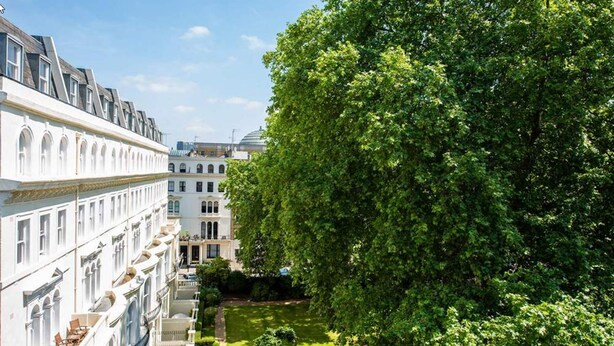 A row of white buildings with arched windows and balconies, a large tree in the foreground, and a well-maintained lawn. - Garden Court Hotel (London)