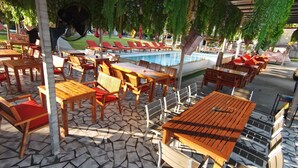 Area piscina all'aperto con tavoli e sedie in legno, poltrona relax e pavimento piastrellato.