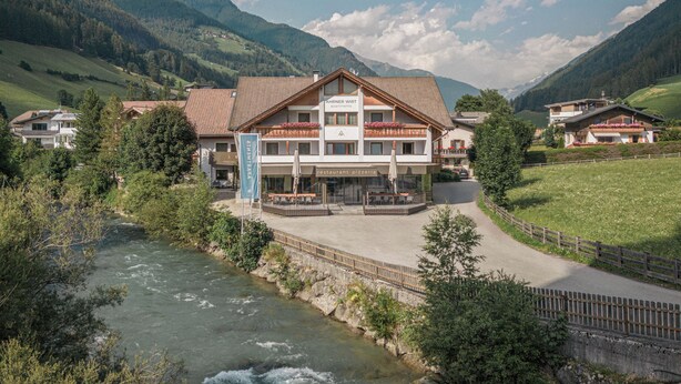 Un hotel con el paisaje de montañas al fondo, un río al frente y un sendero pavimentado.