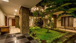 A hotel lobby with a checkered floor, stone pillars, a small garden with tropical plants, and a staircase leading to an upper level. - Svarna Hotel Sanur (Denpasar)