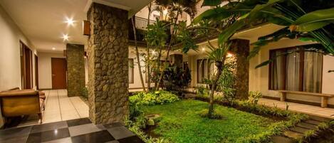 A hotel lobby with a checkered floor, stone pillars, a small garden with tropical plants, and a staircase leading to an upper level.