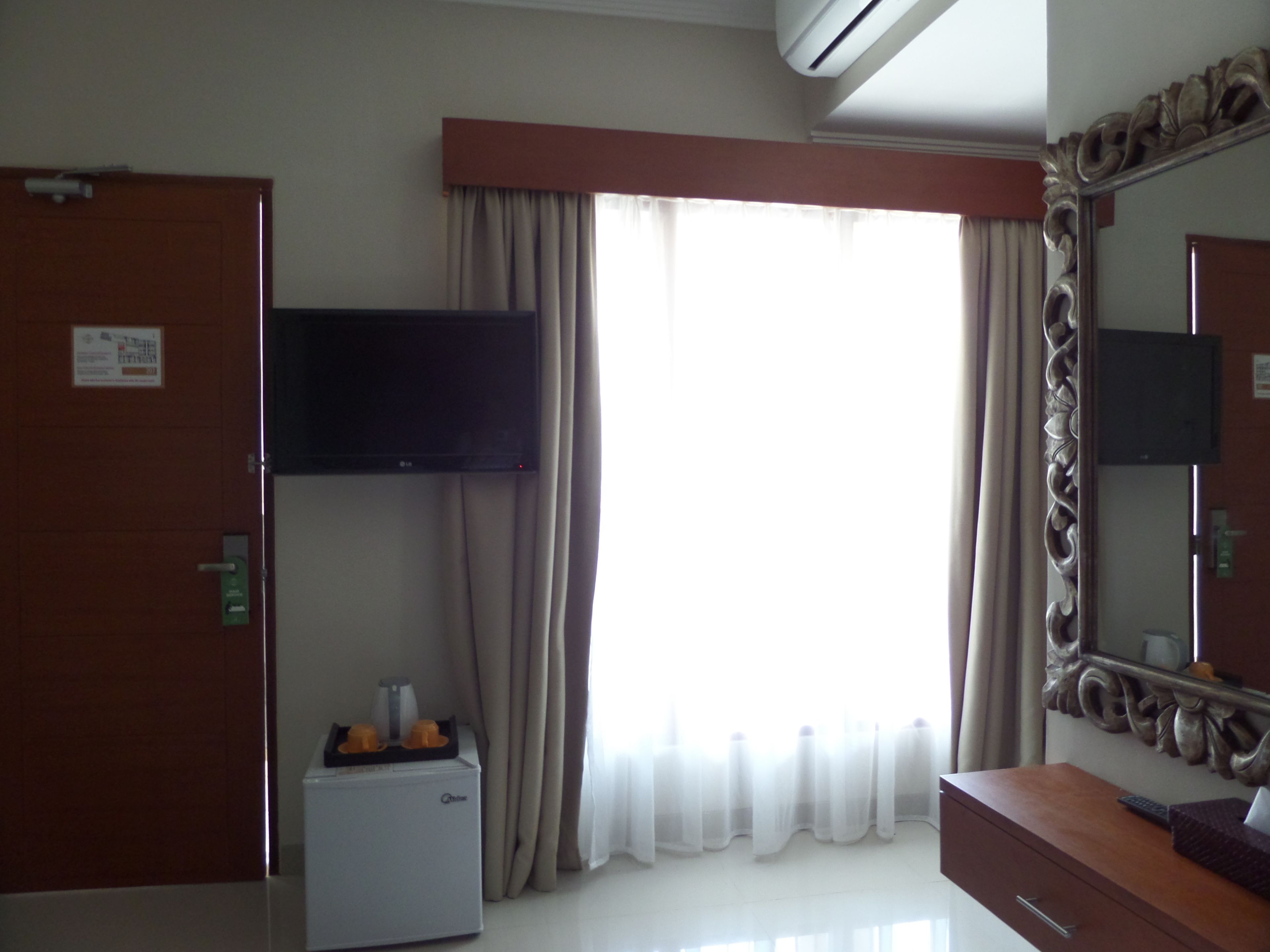 A hotel room with a flat-screen TV mounted on the wall, a mini-fridge, a vanity with a mirror, and a large window with curtains.