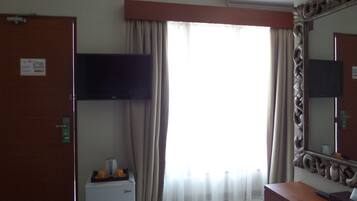 A hotel room with a flat-screen TV mounted on the wall, a mini-fridge, a vanity with a mirror, and a large window with curtains.