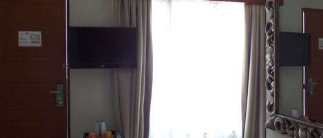 A hotel room with a flat-screen TV mounted on the wall, a mini-fridge, a vanity with a mirror, and a large window with curtains.