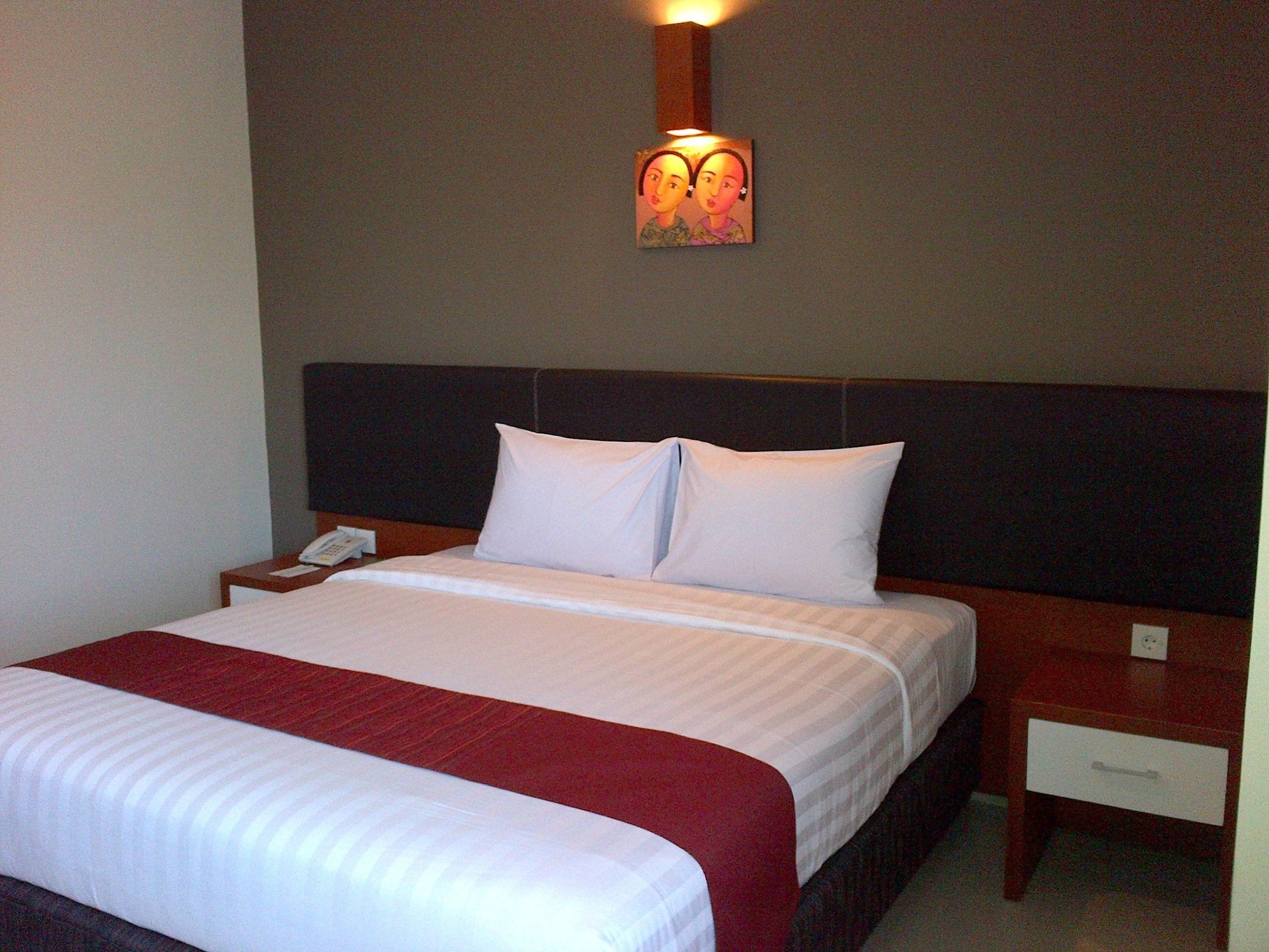 A neatly made bed with white and red bedding, a wooden headboard, a bedside table with a phone, and a wall-mounted light fixture.
