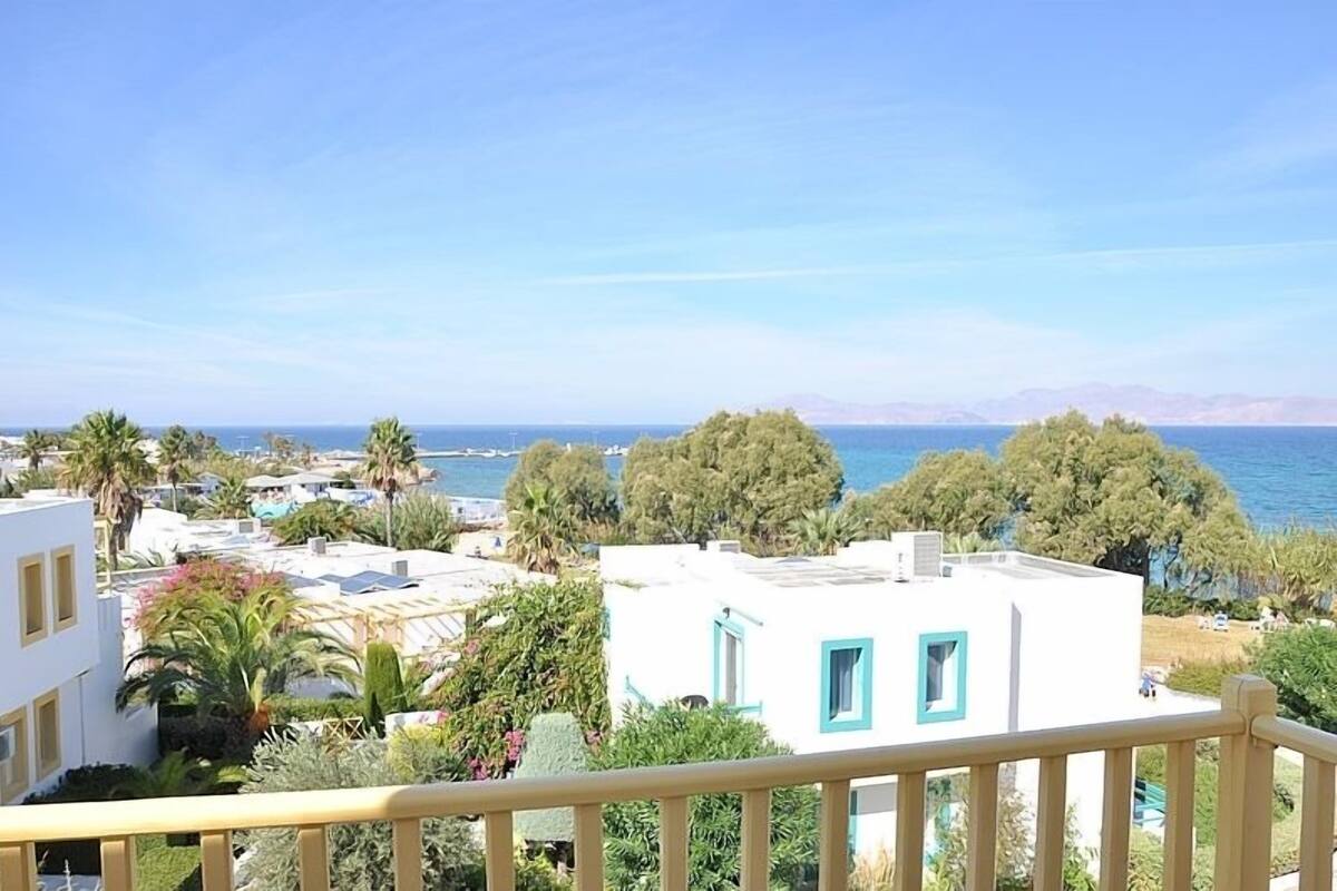 A coastal residential area with white buildings, some with blue trim, and a clear view of the sea and distant mountains.