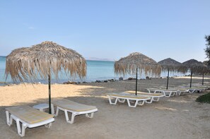 Une plage avec des chaises longues blanches et des parasols en paille, un ciel dégagé et une mer calme.