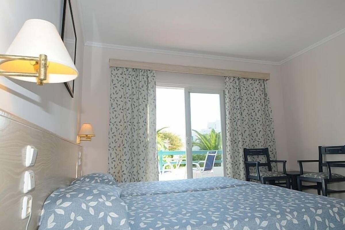 A hotel room with a bed, a desk with chairs, a window with curtains, and a balcony with a chair.