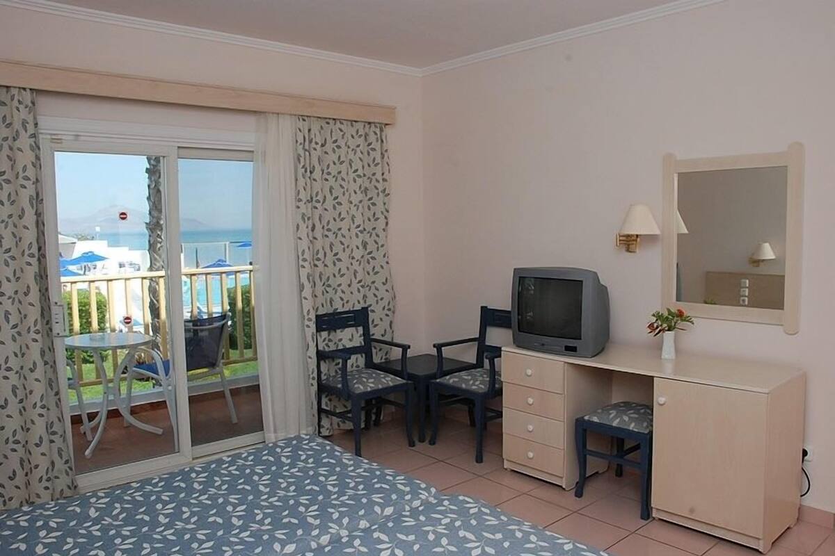 A hotel room with a bed, a desk with a television, a chair, a small stool, a cabinet, and a balcony with a table and chairs.