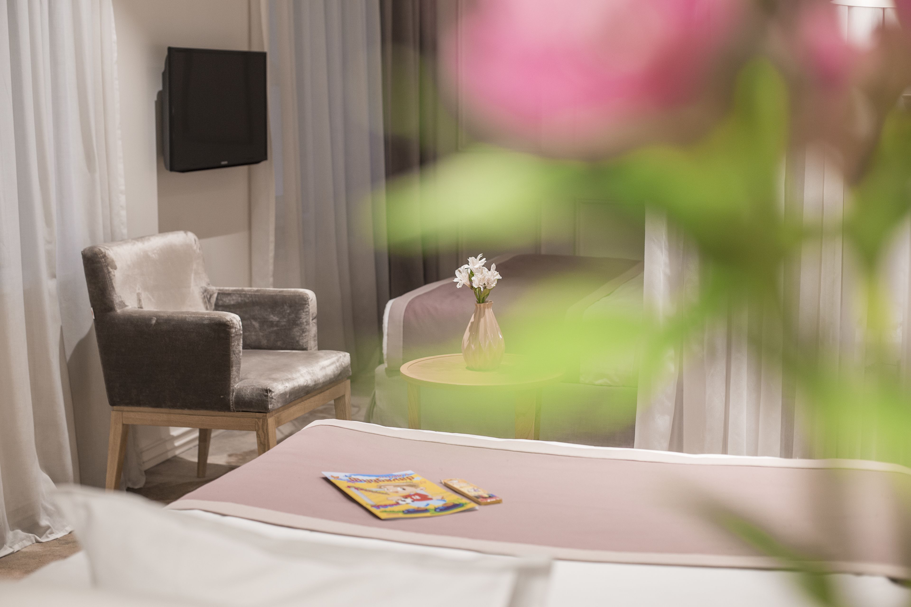 Une pièce comprenant un lit, une chaise, une petite table ronde ornée d’un vase de fleurs et un téléviseur fixé au mur.