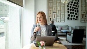 Una donna seduta a un tavolo con un portatile, che tiene in mano un bicchiere d'acqua, in una stanza moderna con una grande finestra e un pannello decorativo.