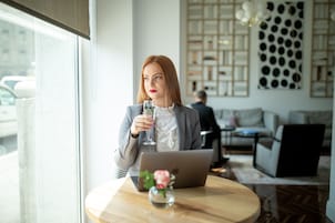 Una mujer sentada a una mesa con una computadora portátil, sosteniendo un vaso de agua, en una habitación moderna con un gran ventanal y un panel decorativo en la pared.